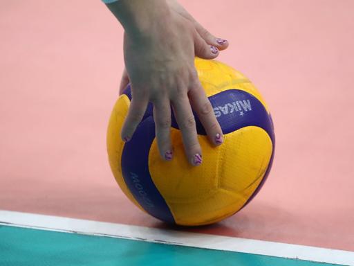 Hände mit lackierten Fingernägel greifen nach einem zweifarbigen Volleyball, der auf dem Hallenboden liegt.