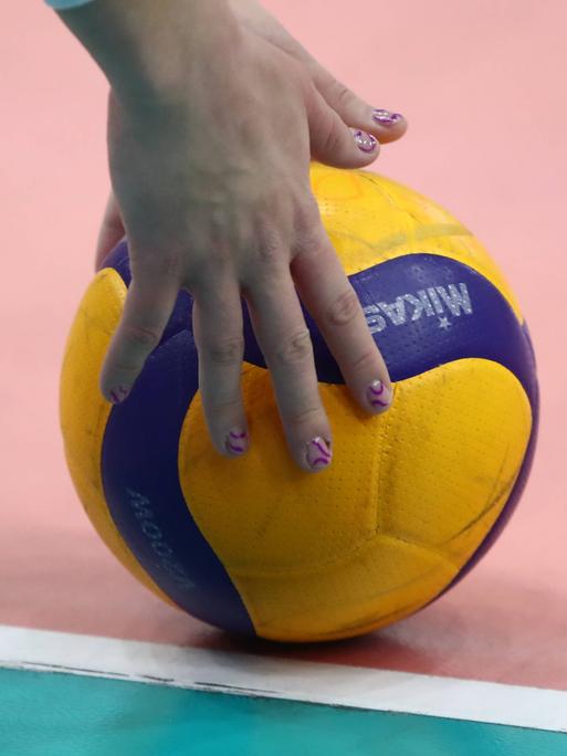 Hände mit lackierten Fingernägel greifen nach einem zweifarbigen Volleyball, der auf dem Hallenboden liegt.