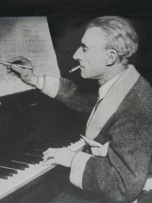 Der Komponist Maurice Ravel sitzt auf einem undatierten Foto am Klavier und hält einen Stift in der rechten Hand, mit dem er auf einem Notenblatt schreibt. 