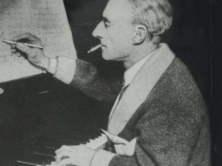 Der Komponist Maurice Ravel sitzt auf einem undatierten Foto am Klavier und hält einen Stift in der rechten Hand, mit dem er auf einem Notenblatt schreibt. 