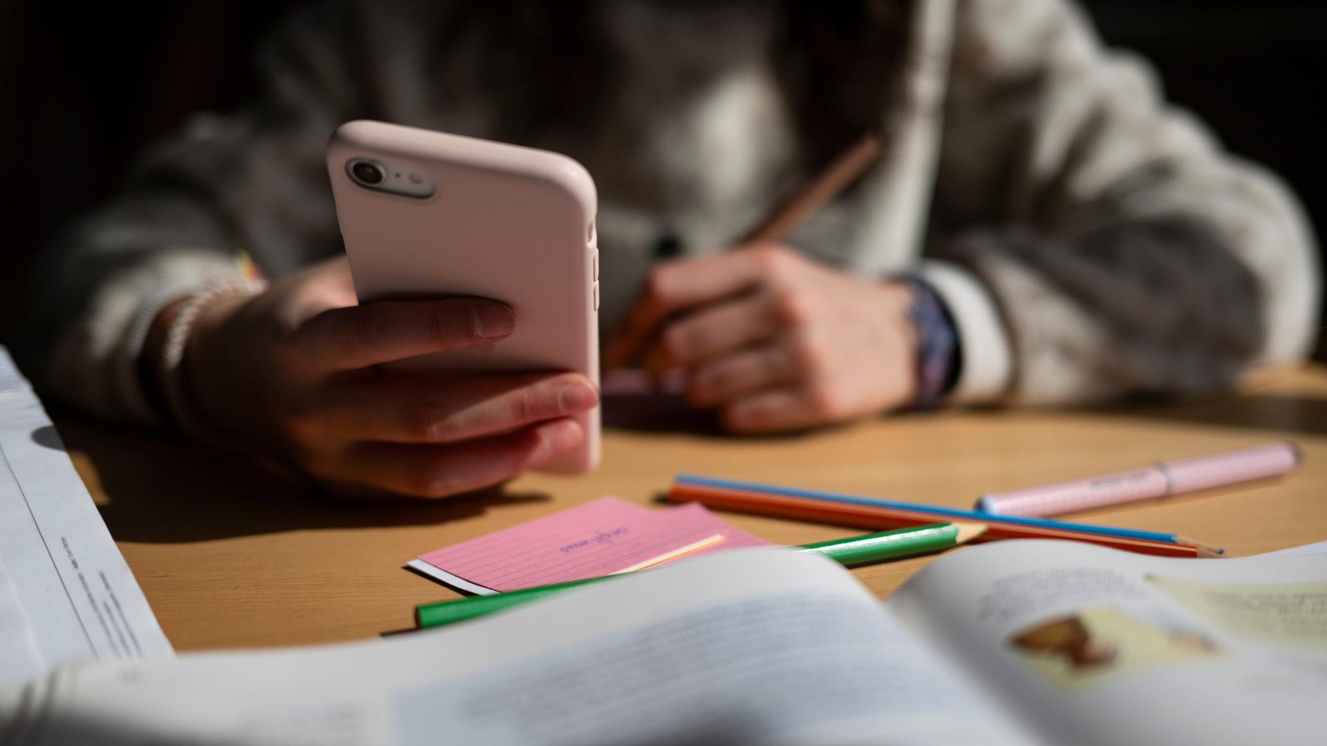 Ein Schulkind sitzt am Schreibtisch, bearbeitet Aufgaben mit einem Stift und hält dabei ein Smartphone in der Hand.