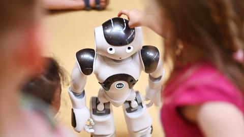Kinder stehen um einen humanoiden Roboter herum. Ein Mädchen tippt ihm auf den Kopf. Kinder stehen um einen humanoiden Roboter herum. Ein Mädchen tippt ihm auf den Kopf.