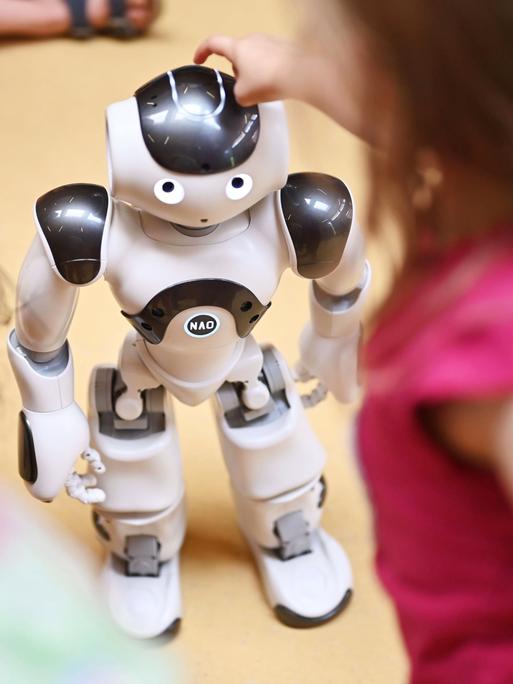 Kinder stehen um einen humanoiden Roboter herum. Ein Mädchen tippt ihm auf den Kopf. Kinder stehen um einen humanoiden Roboter herum. Ein Mädchen tippt ihm auf den Kopf.