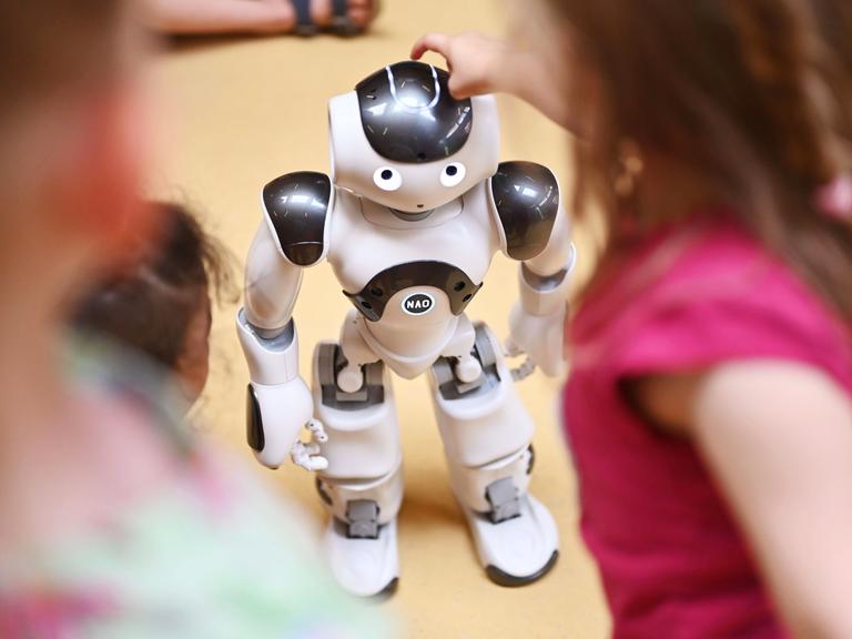 Kinder stehen um einen humanoiden Roboter herum. Ein Mädchen tippt ihm auf den Kopf. Kinder stehen um einen humanoiden Roboter herum. Ein Mädchen tippt ihm auf den Kopf.