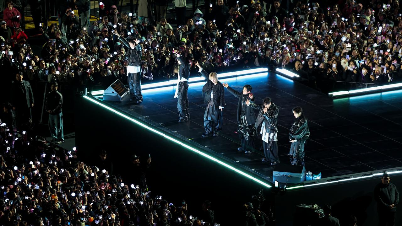 Die Mitglieder von BTS stehen in dunkler Kleidung auf einer Bühne. Sie recken ihre Arme in die Höhe. Rund um die Bühne stehen feiernde Zuschauer.