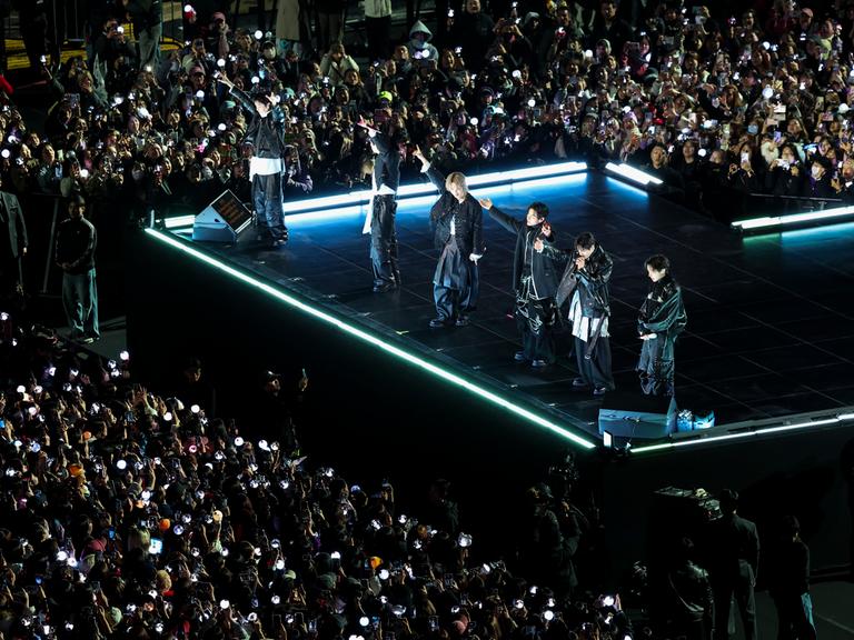 Die Mitglieder von BTS stehen in dunkler Kleidung auf einer Bühne. Sie recken ihre Arme in die Höhe. Rund um die Bühne stehen feiernde Zuschauer.