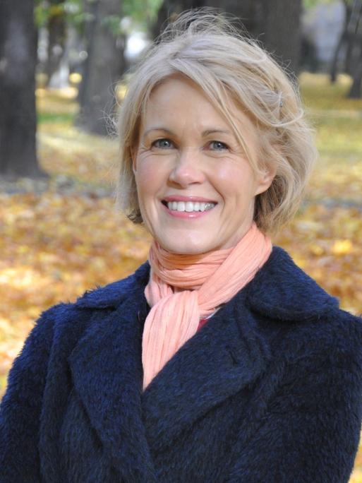 Rósa Lind, eine blondhaarige Frau, steht lächelnd in einem herbstlich anmutenden Park. 