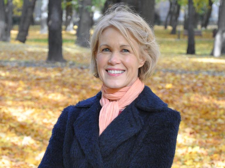 Rósa Lind, eine blondhaarige Frau, steht lächelnd in einem herbstlich anmutenden Park. 