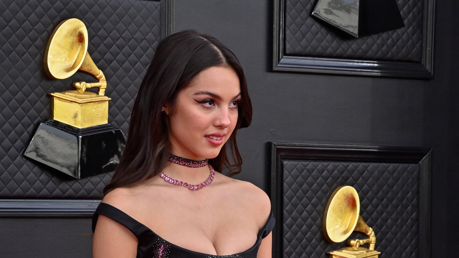 Eine Frau mit schwarzen Haaren und einem offenherzigen Dekolleté steht vor einer Wand, an der Grammy Awards angebracht sind. Eine Frau mit schwarzen Haaren und einem offenherzigen Dekolleté steht vor einer Wand, an der Grammy Awards angebracht sind.