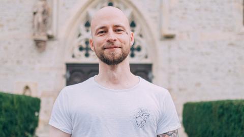 Christopher Schlicht steht vor einer Kirche, trägt ein weißes T-Shirt. Sein linker Arm ist tätowiert.
