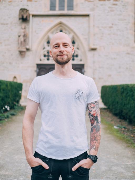 Christopher Schlicht steht vor einer Kirche, trägt ein weißes T-Shirt. Sein linker Arm ist tätowiert.