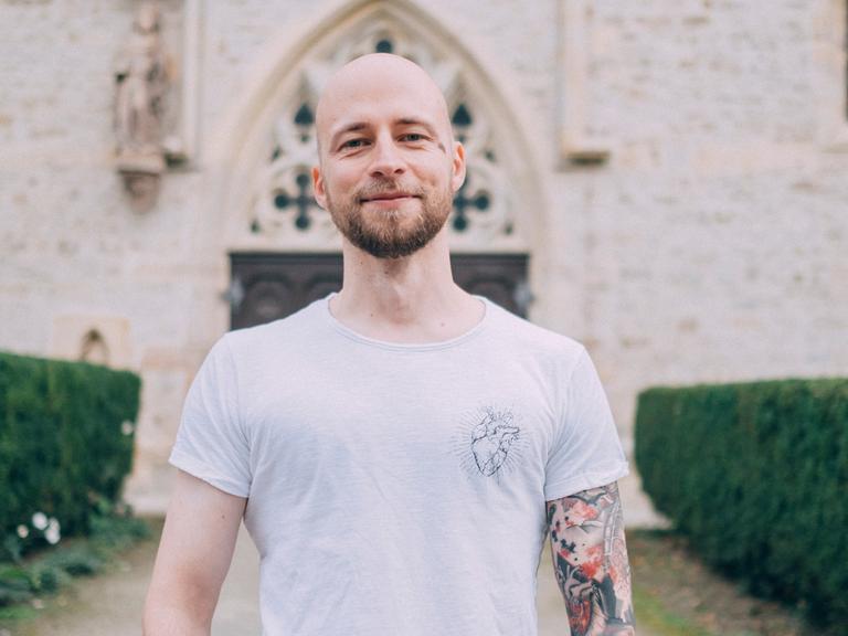 Christopher Schlicht steht vor einer Kirche, trägt ein weißes T-Shirt. Sein linker Arm ist tätowiert.