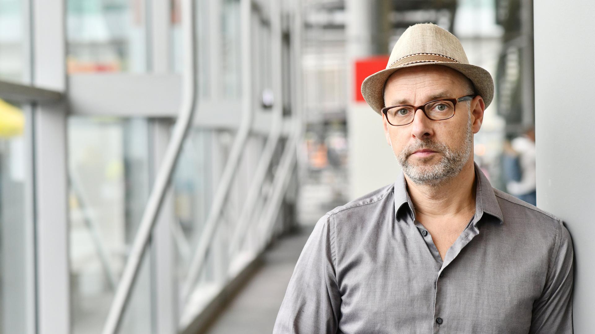 Comiczeichner Reinhard Kleist steht mit einem Hut auf dem Kopf an eine Wand gelehnt. Im Hintergrund ein gläserner Flur auf der Buchmesse in Frankfurt am Main. 