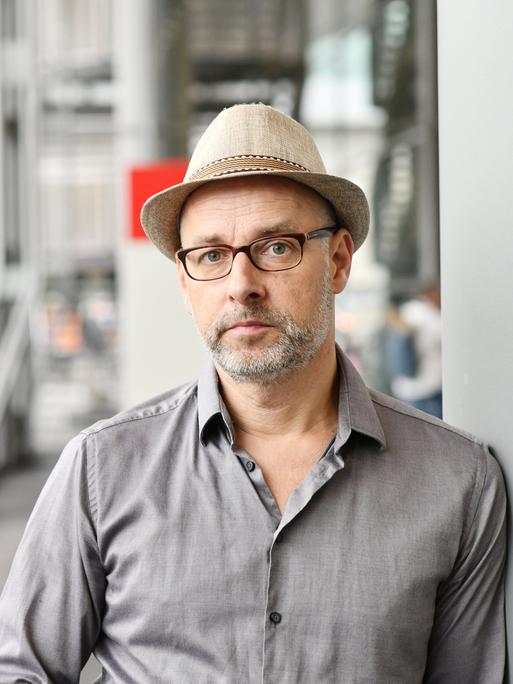 Comiczeichner Reinhard Kleist steht mit einem Hut auf dem Kopf an eine Wand gelehnt. Im Hintergrund ein gläserner Flur auf der Buchmesse in Frankfurt am Main. 