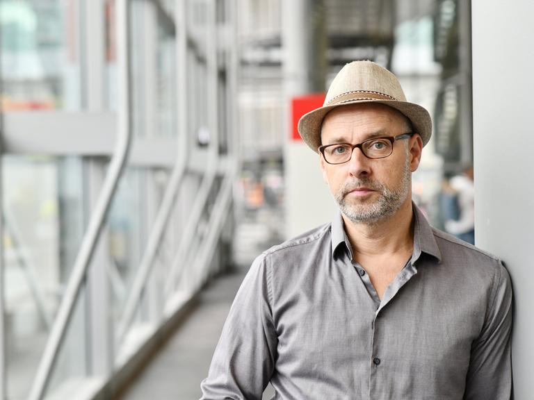 Comiczeichner Reinhard Kleist steht mit einem Hut auf dem Kopf an eine Wand gelehnt. Im Hintergrund ein gläserner Flur auf der Buchmesse in Frankfurt am Main. 