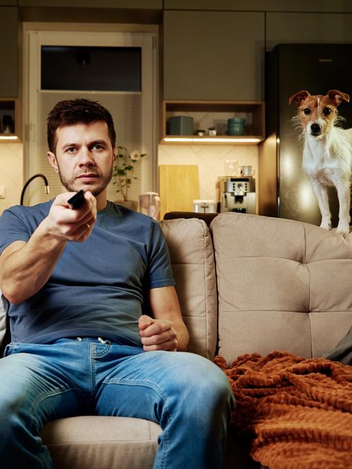Ein Mann sitzt zu Hause auf dem Sofa und schaut Richtung Kamera; er hält eine Fernbedienung in der Hand. Auf dem Sofa steht ein Hund, der ebenfalls in Richtung Kamera schaut.