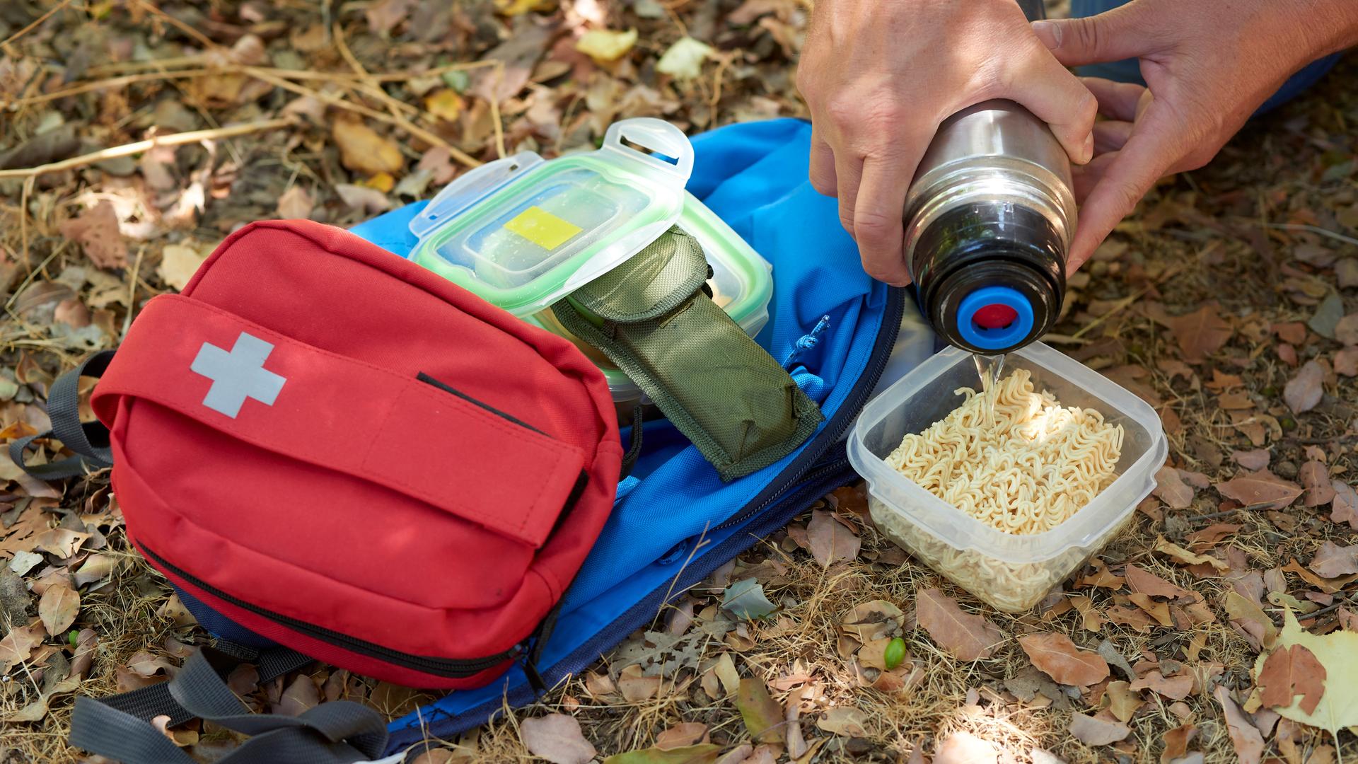 Nahaufnahme einer Szene in der sich jemand neben einem Notfallrucksack mit heißem Wasser aus einer Thermoskanne und Instantnudeln eine Mahlzeit zubereitet. Daneben liegt ein Erste-Hilfe-Set. Nahaufnahme einer Szene in der sich jemand neben einem Notfallrucksack mit heißem Wasser aus einer Thermoskanne und Instantnudeln eine Mahlzeit zubereitet. Daneben liegt ein Erste-Hilfe-Set.
