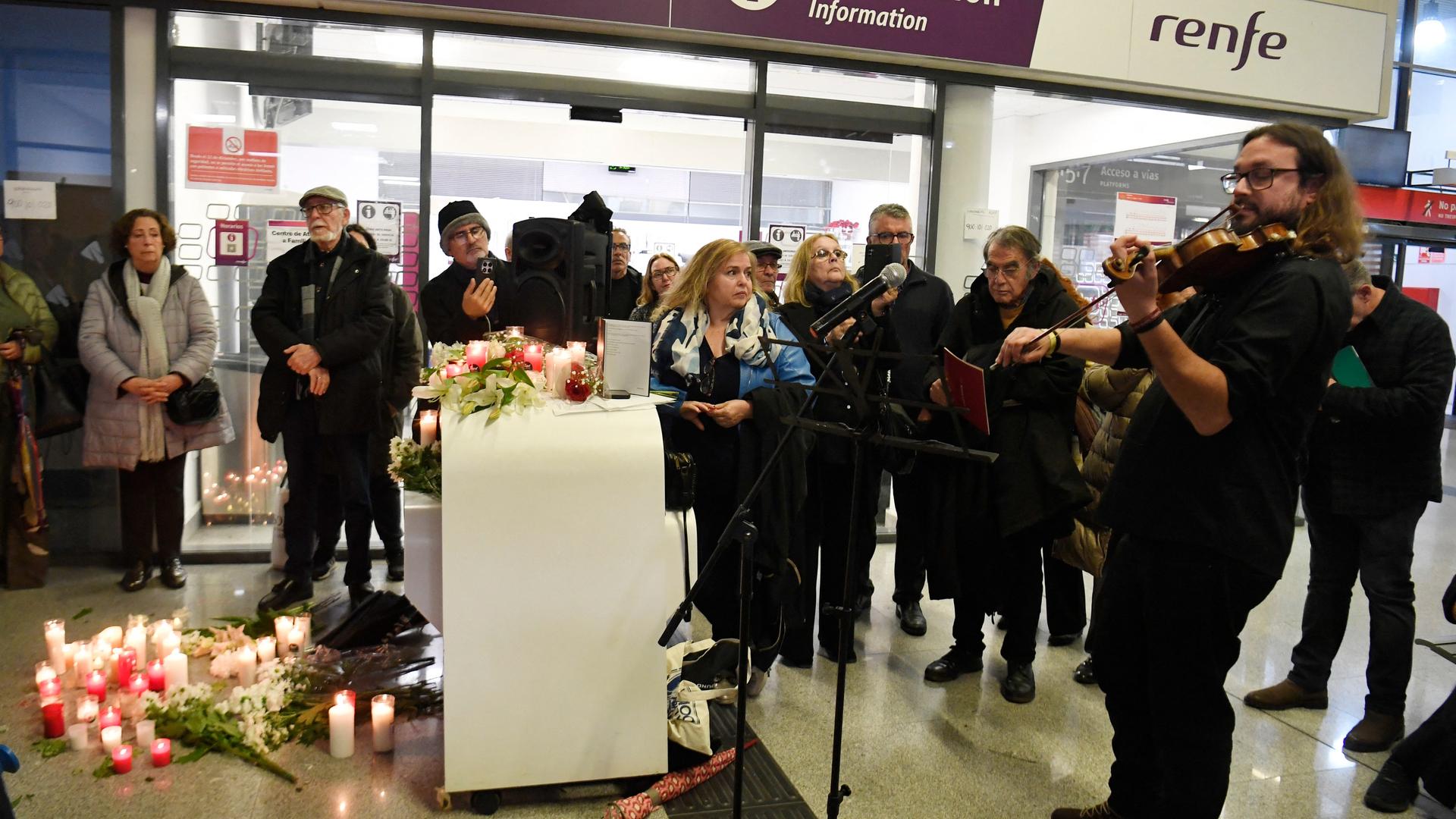 Menschen stehen im Bahnhof von Huelva um Blumen und Kerzen versammelt. Es spielt ein Mann auf einer Geige. Menschen stehen im Bahnhof von Huelva um Blumen und Kerzen versammelt. Es spielt ein Mann auf einer Geige.