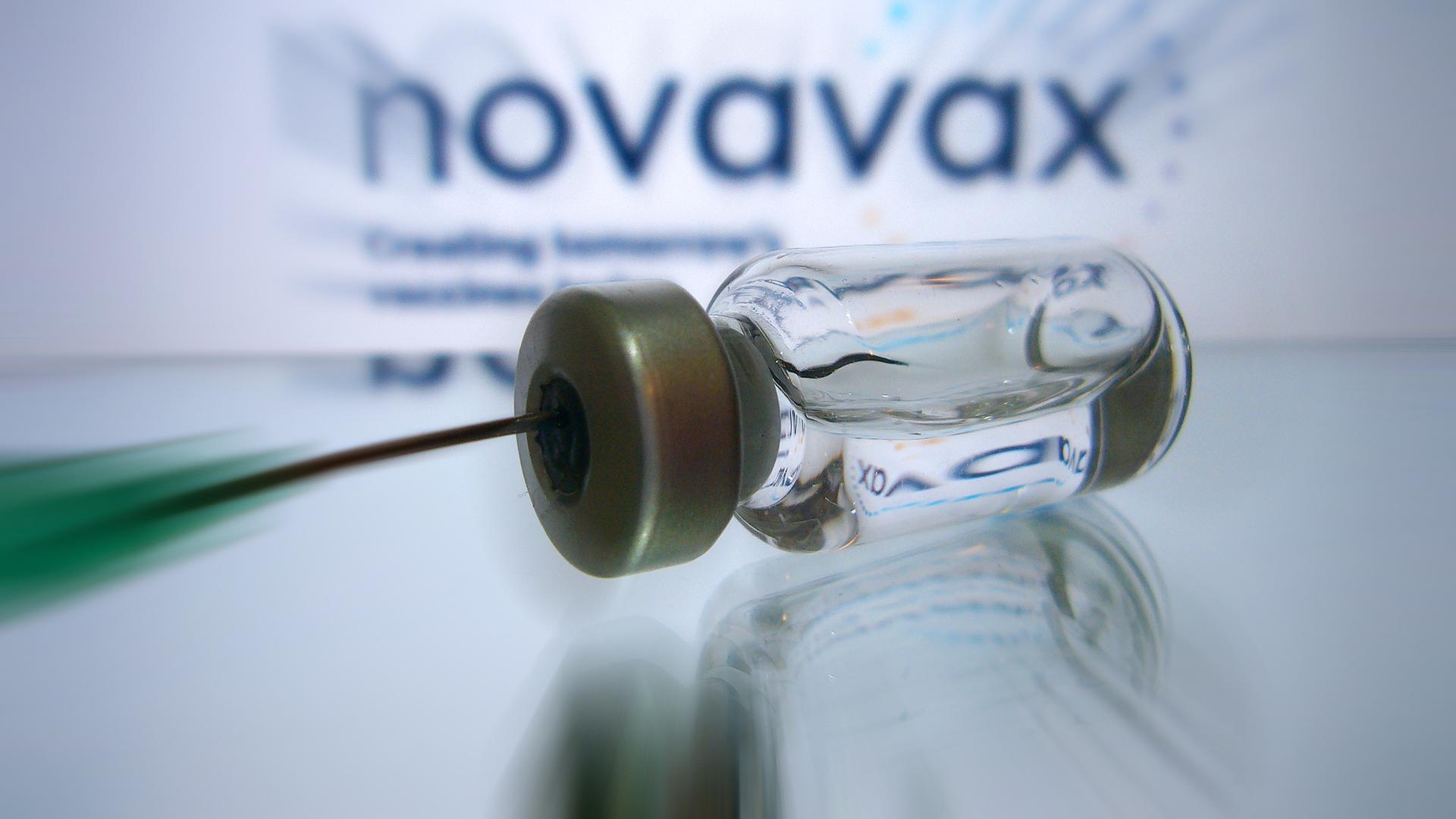 Eine Kanüle zieht eine Impfdosis aus einen Fläschchen auf. Im Hintergrund: das Logo von Novavax (Symbolfoto) 