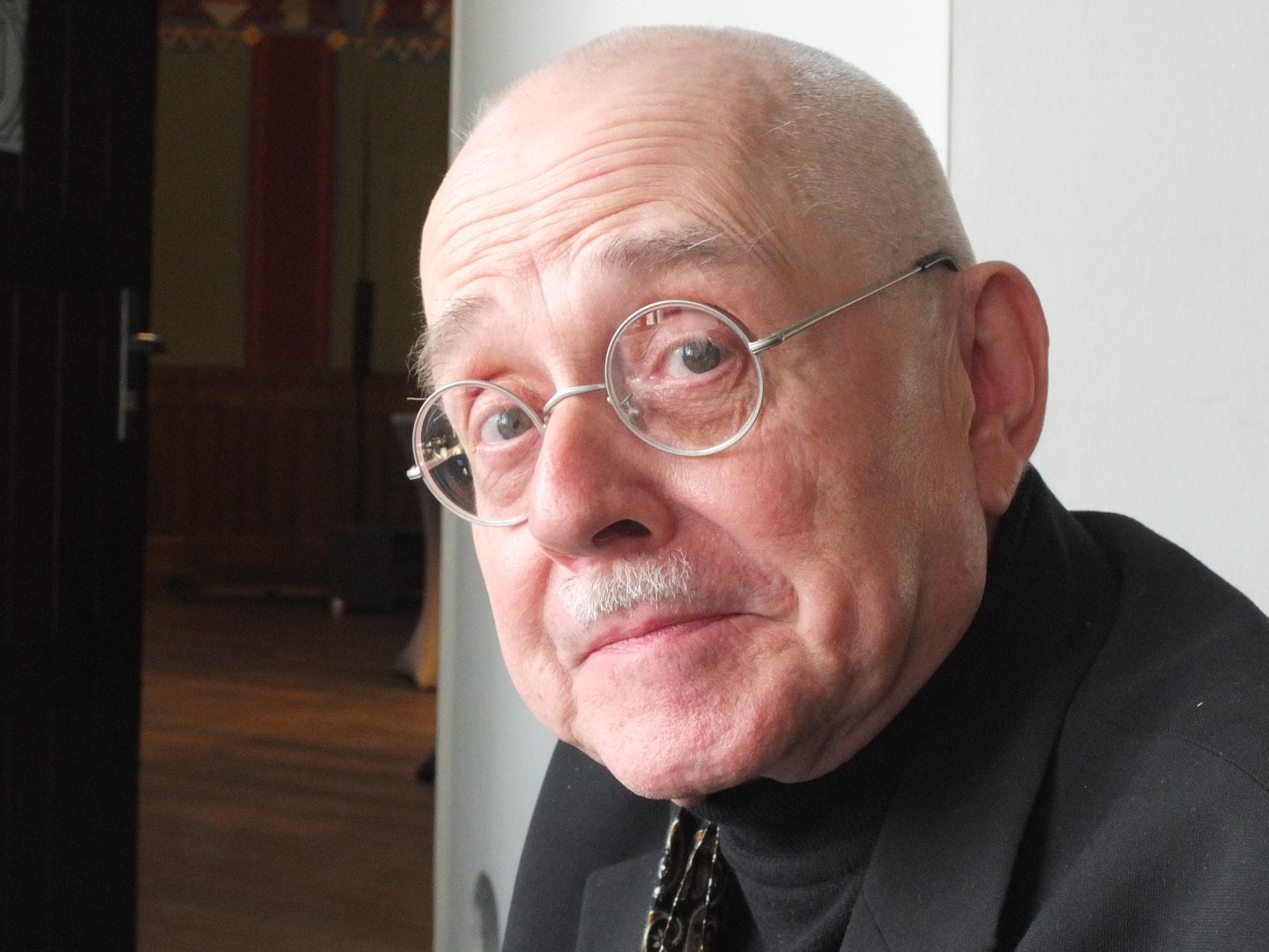 Ein älterer Mann mit runder Brille sitzt vor einer geöffneten Tür und blickt in die Kamera