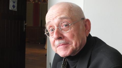 Ein älterer Mann mit runder Brille sitzt vor einer geöffneten Tür und blickt in die Kamera