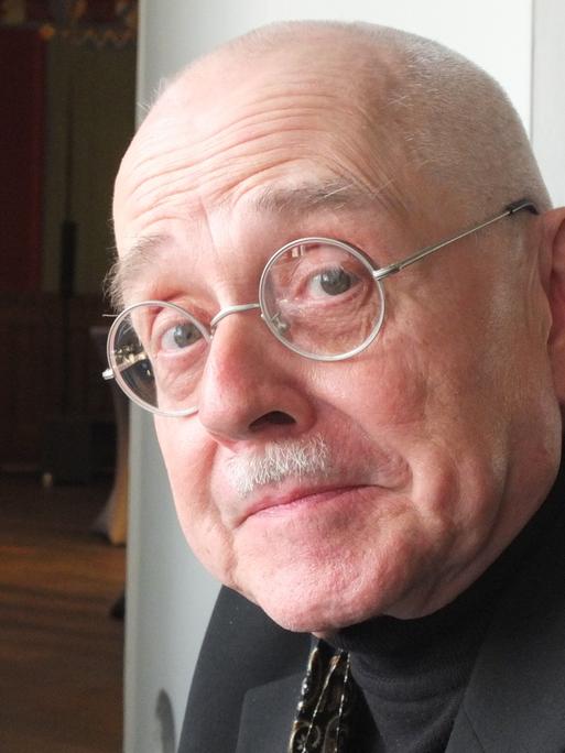 Ein älterer Mann mit runder Brille sitzt vor einer geöffneten Tür und blickt in die Kamera