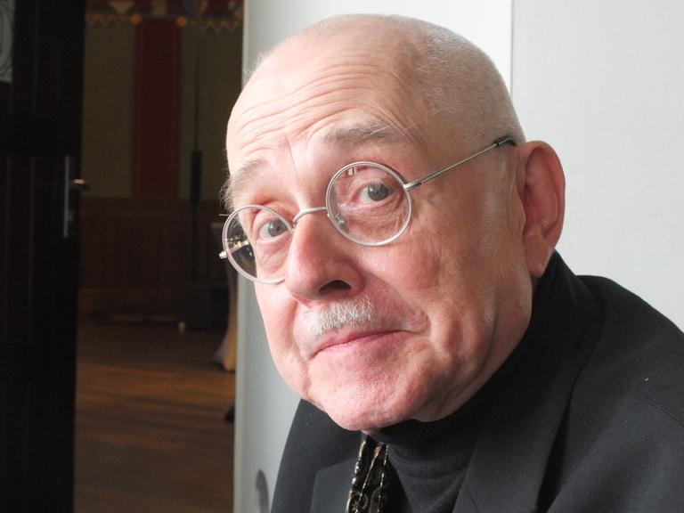 Ein älterer Mann mit runder Brille sitzt vor einer geöffneten Tür und blickt in die Kamera