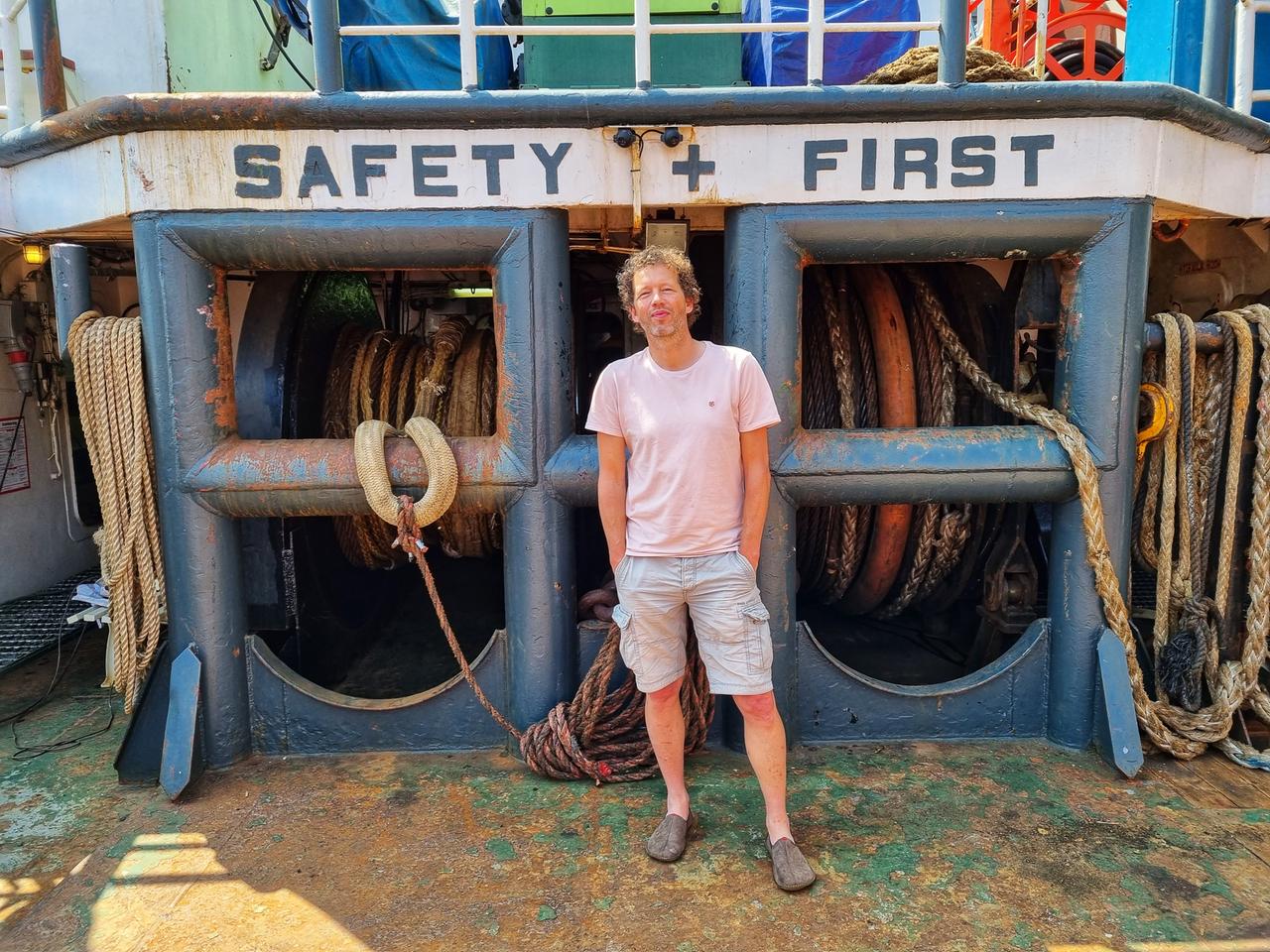 Algenfarmer Joost Wouters aus den Niederlanden steht auf einem Boot an der Küste. Er trägt kurze Hose und T-Shirt. Die Sonne scheint.