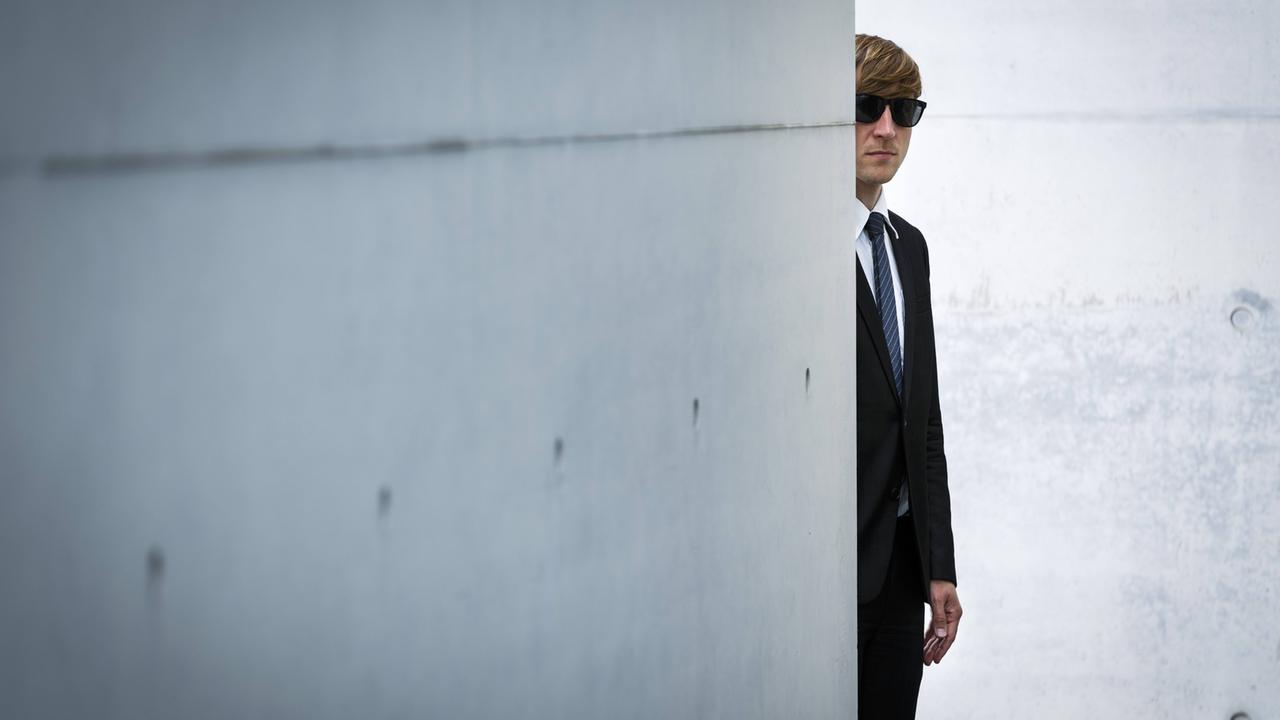 Ein Mann mit Anzug und Sonnenbrille versteckt sich hinter einer Betonmauer. Ein Mann mit Anzug und Sonnenbrille versteckt sich hinter einer Betonmauer.