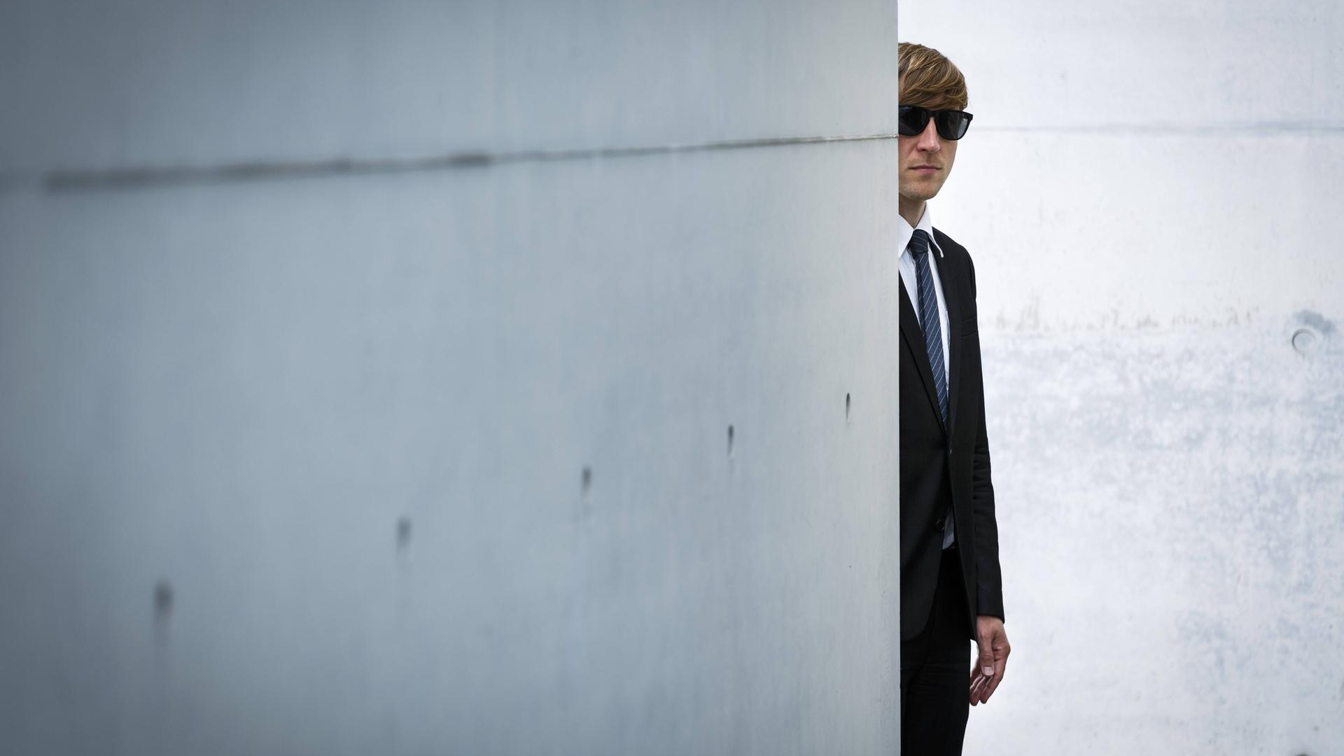 Ein Mann mit Anzug und Sonnenbrille versteckt sich hinter einer Betonmauer.