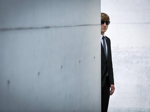 Ein Mann mit Anzug und Sonnenbrille versteckt sich hinter einer Betonmauer. Ein Mann mit Anzug und Sonnenbrille versteckt sich hinter einer Betonmauer.