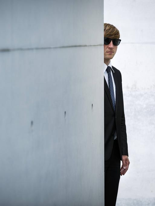 Ein Mann mit Anzug und Sonnenbrille versteckt sich hinter einer Betonmauer.