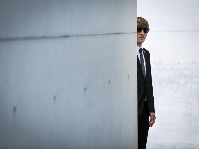 Ein Mann mit Anzug und Sonnenbrille versteckt sich hinter einer Betonmauer.