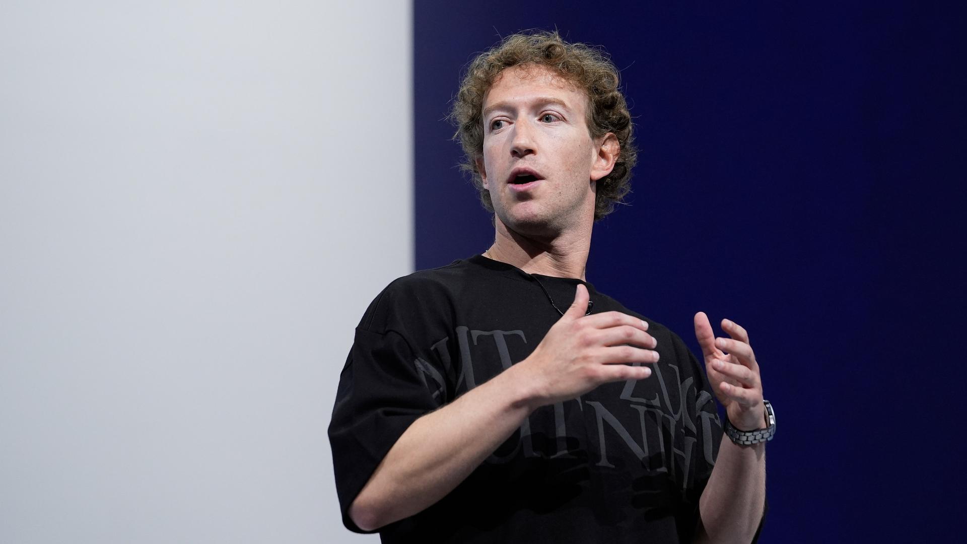 Mark Zuckerberg spricht auf einer Konferenz vor einem blau-weißen Hintergrund. Er trägt ein schwarzes T-Shirt mit grauer Schrift und gestikuliert. 