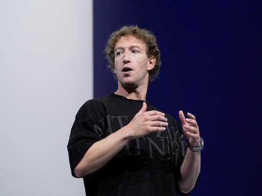 Mark Zuckerberg spricht auf einer Konferenz vor einem blau-weißen Hintergrund. Er trägt ein schwarzes T-Shirt mit grauer Schrift und gestikuliert. 