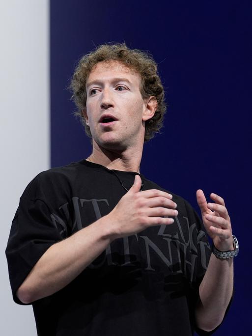 Mark Zuckerberg spricht auf einer Konferenz vor einem blau-weißen Hintergrund. Er trägt ein schwarzes T-Shirt mit grauer Schrift und gestikuliert. 