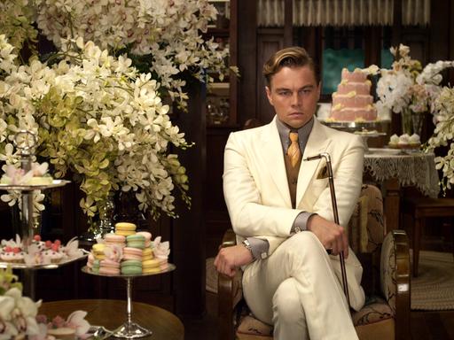 Szene aus dem Film "Der große Gatsby" mit Leonardo DiCaprio in der Titelrolle. Er sitzt in einem weißen Anzug zwischen lauter Blumengestecken und Gedecken mit Törtchen und hält einen Gehstock in der Hand. Dabei schaut er sehr nachdenklich.