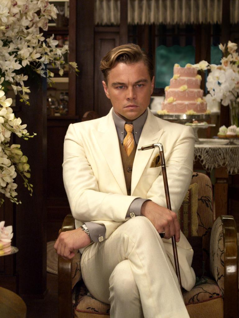 Szene aus dem Film "Der große Gatsby" mit Leonardo DiCaprio in der Titelrolle. Er sitzt in einem weißen Anzug zwischen lauter Blumengestecken und Gedecken mit Törtchen und hält einen Gehstock in der Hand. Dabei schaut er sehr nachdenklich.