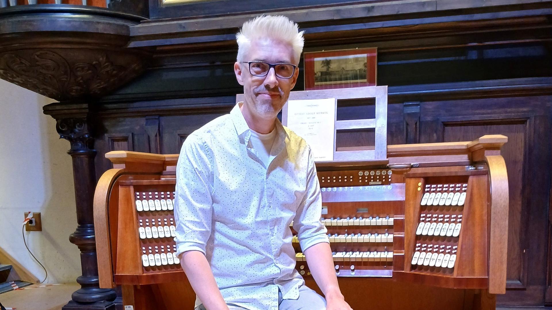 Ein Organist mit blonden, kurzen Haaren sitzt auf einer Orgelbank. Hinter ihm sind übereinander liegende Tastenreihen und Registerzüge zu sehen.