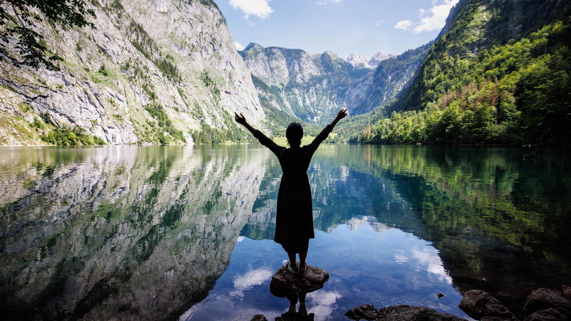 Eine junge Frau steht auf einem Stein im Wasser vom Ober-See und blickt auf das umliegende Berg-Panorama im National-Park Berchtesgaden. Eine junge Frau steht auf einem Stein im Wasser vom Ober-See und blickt auf das umliegende Berg-Panorama im National-Park Berchtesgaden.