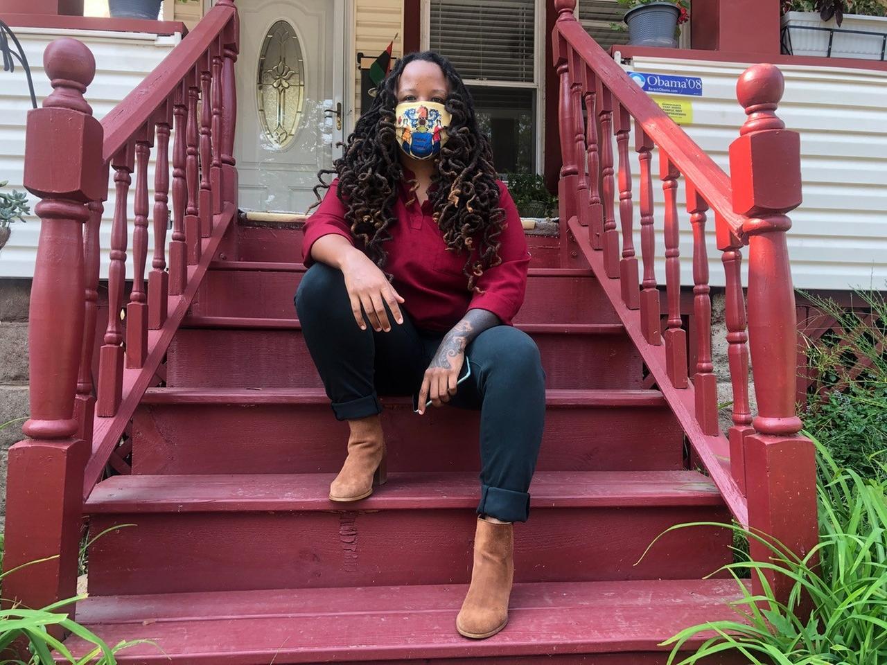 Imani Oakley sitzt auf der Treppe ihres Wohnhauses in der Stadt Montclair. Sie hat lange, schwarze Haare und trägt einen bunten Mund-Nasen-Schutz im Gesicht.