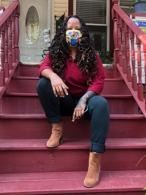 Imani Oakley sitzt auf der Treppe ihres Wohnhauses in der Stadt Montclair. Sie hat lange, schwarze Haare und trägt einen bunten Mund-Nasen-Schutz im Gesicht.