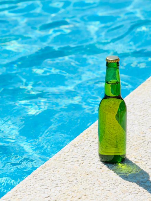 Eine kühle, grüne Flasche Bier steht am Rande eines tiefblauen Pools in der Sonne. Syymbolbild für Urlaub. 
