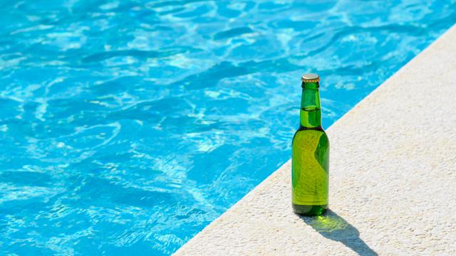 Eine grüne Flasche Bier steht am Rande eines tiefblauen Pools in der Sonne. 