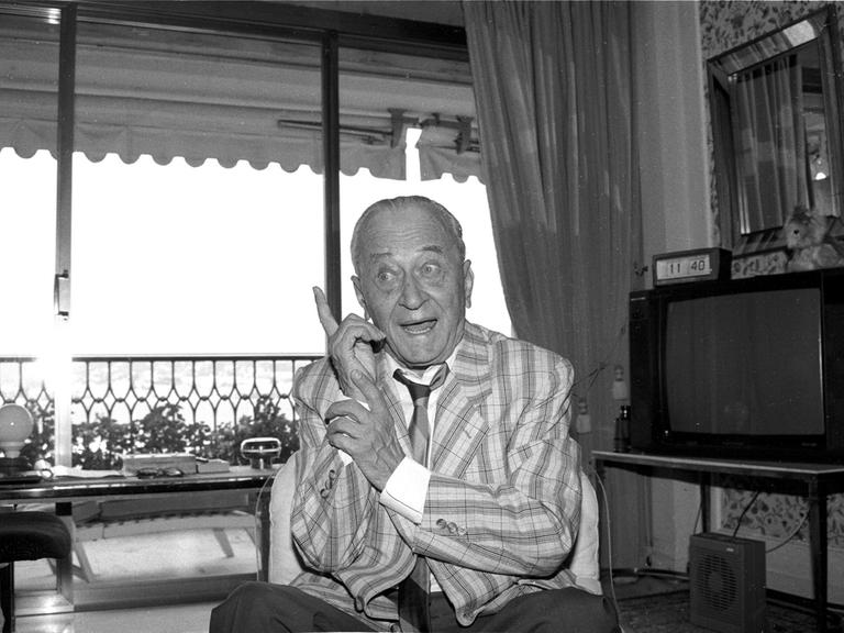 Der Journalist, Erfinder und Medienmanager Eduard Rhein Eduard Rhein sitzt in Jacket, Hemd und Krawatte in einem Wohnzimmern. Lächelnd hebt er den Zeigefiner der linken Hand. Der Journalist, Erfinder und Medienmanager Eduard Rhein Eduard Rhein sitzt in Jacket, Hemd und Krawatte in einem Wohnzimmern. Lächelnd hebt er den Zeigefiner der linken Hand.