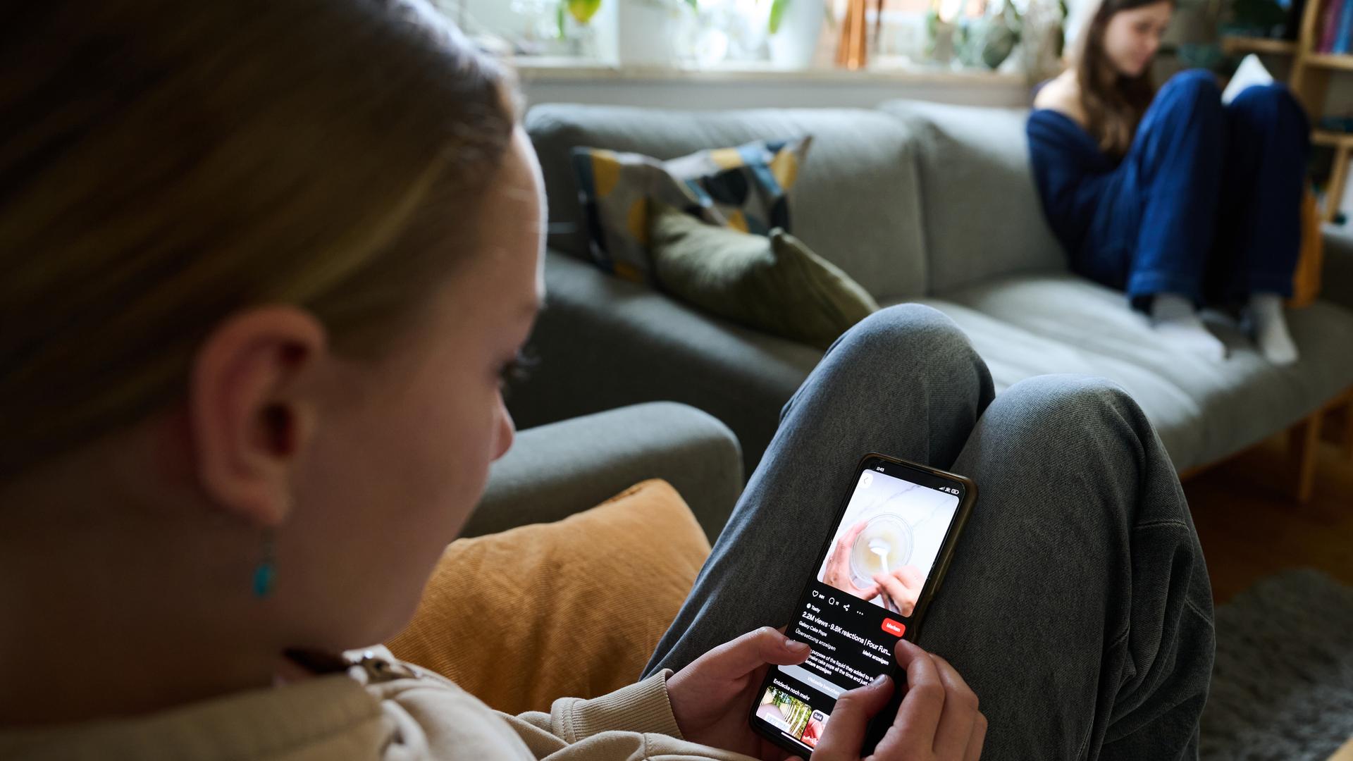 Ein Mädchen blickt auf sein Smartphone, während ein anderes Mädchen ein Buch liest.