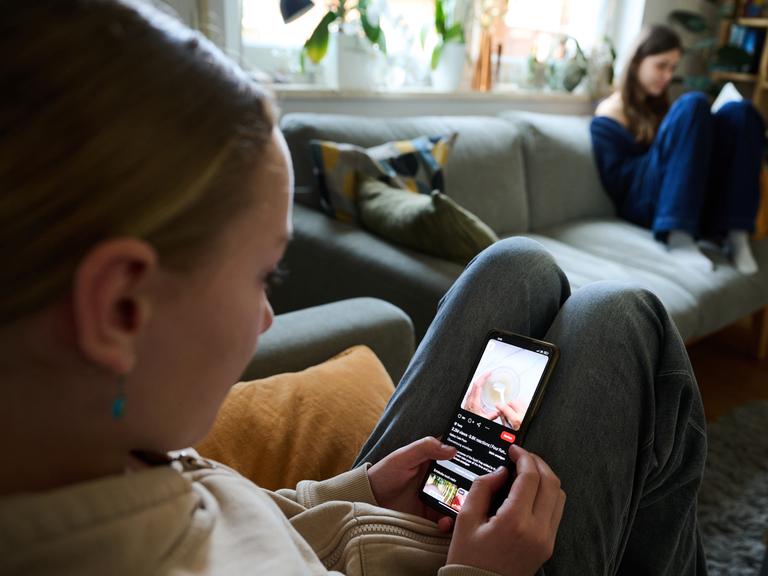 Ein Mädchen blickt auf sein Smartphone, während ein anderes Mädchen ein Buch liest.