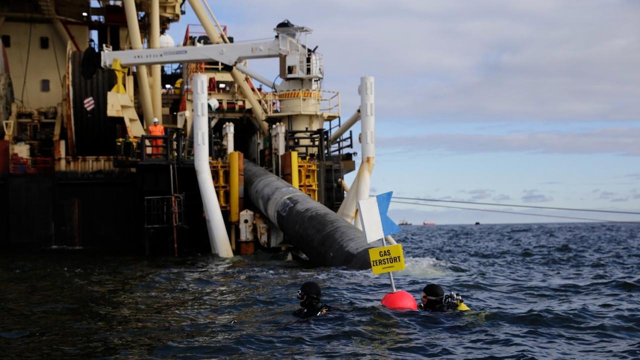 Zu sehen ist das Pipeline-Verlegeschiff "Castoro 10", davor zwei Taucher im Wasser und eine Boje mit einem Schild, auf dem steht "Gas zerstört".