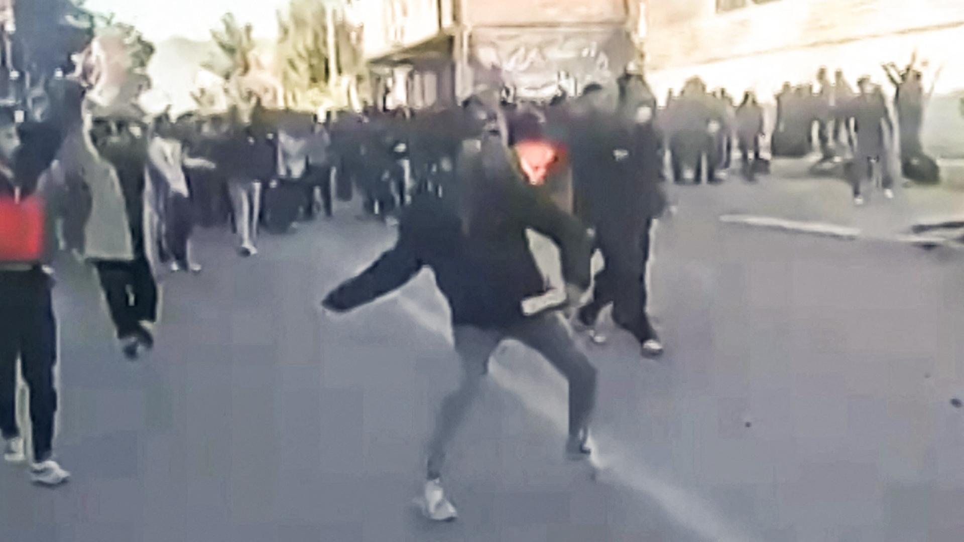 Der verpixelte Videoscreenshot zeigt eine demonstrierende Menschenmenge. Einzelne Personen im Vordergrund werfen mit Steinen.