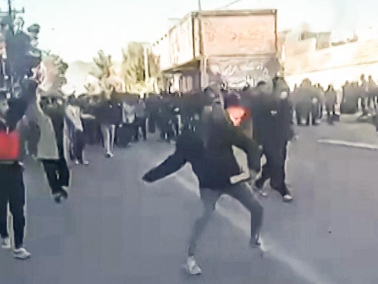 Der verpixelte Videoscreenshot zeigt eine demonstrierende Menschenmenge. Einzelne Personen im Vordergrund werfen mit Steinen.